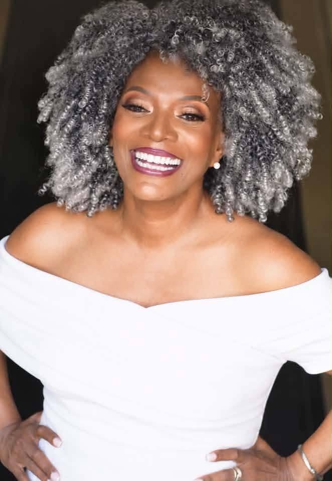 A confident, smiling woman with silver curls in a white off-shoulder dress